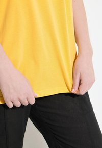Žluté bavlněné tričko s kulatým výstřihem, volným střihem a krátkými rukávy, spárované s černými kalhotami. Hladká textura, minimální detaily.