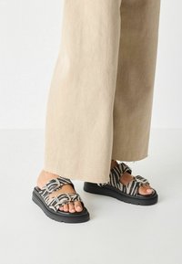 Sandales compensées noires avec des brides imprimé zèbre et des boucles ajustables, associées à un pantalon beige à jambes larges. Matériau texturé, design décontracté.