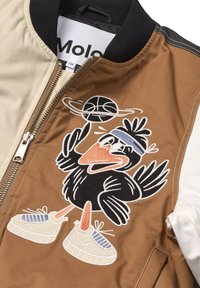 Brun og beige jakke med sort krave, der har et broderet tegneserie-fugl, der spinner en basketball iført et svedbånd og sneakers.