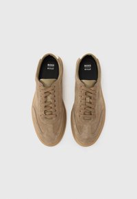 Brown suede sneakers with round toe, tonal laces, beige accents on heel, and a textured rubber sole. Black insole with brand name.