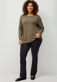 Maglietta verde oliva a maniche lunghe con scollatura tonda e vestibilità rilassata, abbinata a pantaloni neri aderenti e scarpe basse nere.