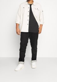 Camicia bianca con bottoni e maniche lunghe sopra una t-shirt nera, abbinata a pantaloni neri e sneaker bianche, con le maniche arrotolate.