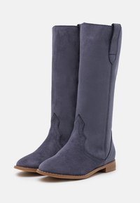 Para wysokich, granatowych zamszowych botków z wąskimi noskami, ozdobnym przeszyciem, uchwytami do zakładania i niskimi, brązowymi płaskimi obcasami na białym tle.