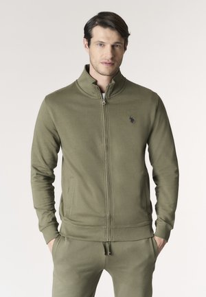 Uomo che indossa una giacca con zip color verde oliva e pantaloni abbinati, in piedi con le mani nelle tasche su uno sfondo semplice.