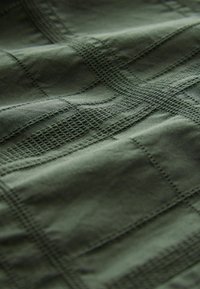 Groene stof met een getextureerd patroon met rasterachtige lijnen en gestikte accenten; gladde textuur met een subtiele glans.