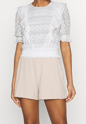 Le modèle porte un chemisier blanc en dentelle à manches bouffantes et à l’ourlet festonné, assorti à un short plissé beige.