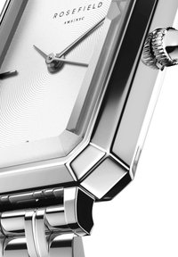 Srebrny prostokątny zegarek z teksturowaną białą tarczą, cienkimi srebrnymi wskazówkami i wyraźnym koroną. Polerowana metalowa obudowa z kanciastymi detalami.