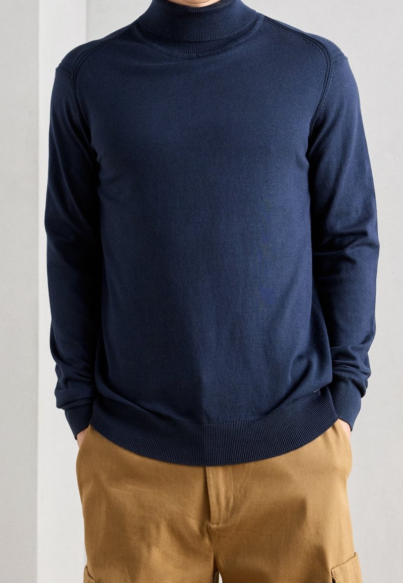 Dunkelblauer gestrickter Rollkragenpullover mit gerippten Bündchen und Saum, kombiniert mit beigen Cargo-Hosen, die eine glatte Textur aufweisen.