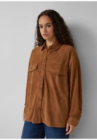 Junge Frau mit langen lockigen Haaren, die ein lockeres braunes Hemd mit Knöpfen und blaue Jeans trägt, steht an einer grauen Wand.