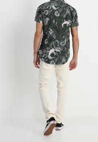 Zielona koszula z krótkim rękawem w biały wzór paisley; zestawiona z jasnobeżowymi dżinsami i czarnymi sneakersami. Strukturalny krój i swobodny styl.