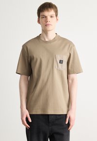 Tricou scurt cu mânecă scurtă, de culoare bej, cu guler rotund, având o buzunar texturat și un patch negru al mărcii pe partea stângă. Material din bumbac.