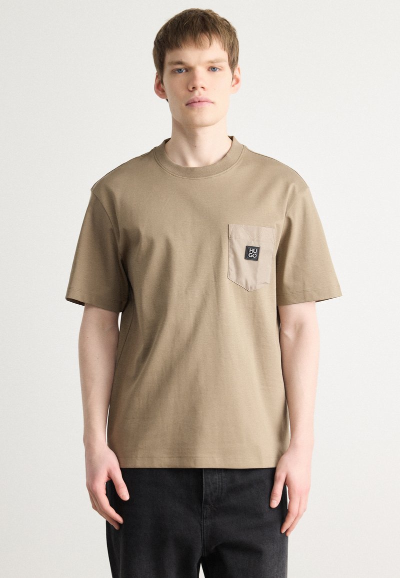 Tricou scurt cu mânecă scurtă, de culoare bej, cu guler rotund, având o buzunar texturat și un patch negru al mărcii pe partea stângă. Material din bumbac.