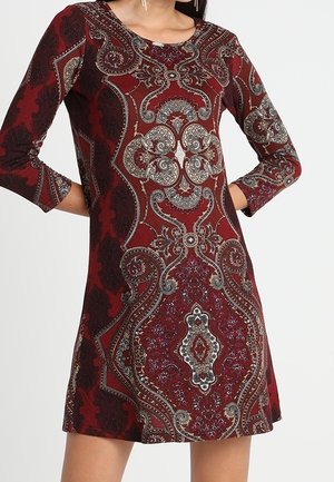 Kobieta ubrana w czerwoną sukienkę z długim rękawem z misternym wzorem paisley i ozdobnym, stojąca z rękami na biodrach na jednolitym tle.