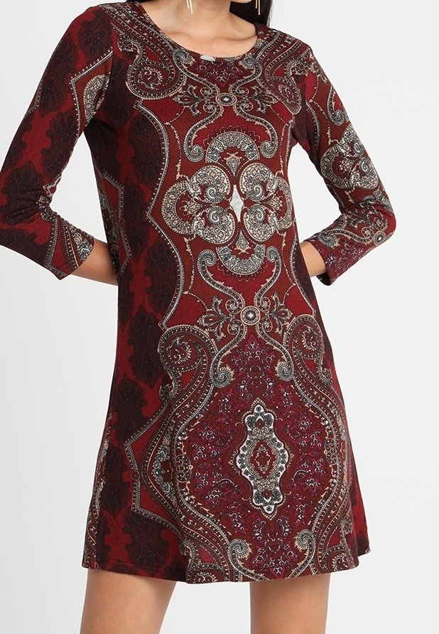 Femme portant une robe rouge à manches longues avec un motif complexe de paisley et ornemental, debout les mains sur les hanches devant un fond uni.