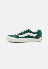 Zapatilla de ante verde con detalles en blanco, punta redondeada y cordones planos. Presenta un diseño clásico con suela de goma y detalle de franja negra.