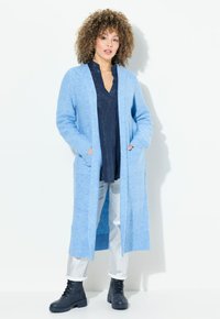 Ljusblå lång cardigan i mjukt, strukturerat stickat material. Har två framfickor och ribbade muddar. Bärs över en mörk topp och silverfärgade byxor.