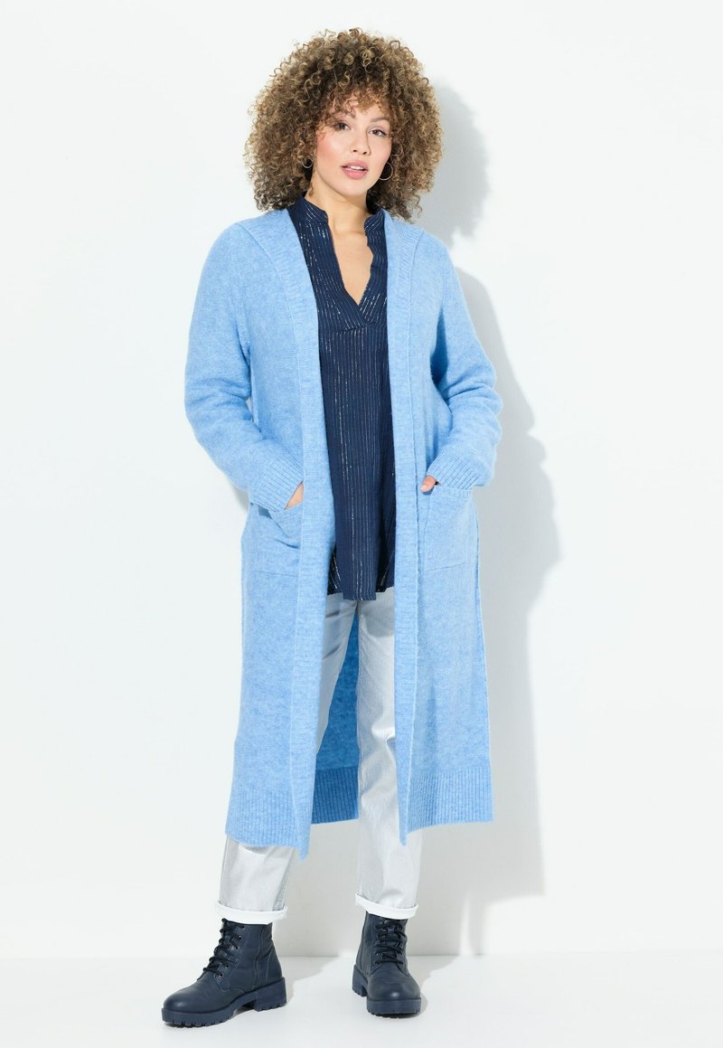 Ljusblå lång cardigan i mjukt, strukturerat stickat material. Har två framfickor och ribbade muddar. Bärs över en mörk topp och silverfärgade byxor.