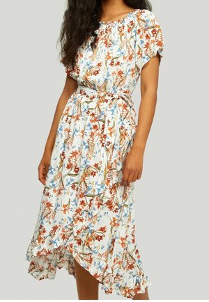 Donna che indossa un vestito bianco a fiori con motivi arancioni, blu e verdi, maniche corte, cintura in vita e orlo asimmetrico arricciato.