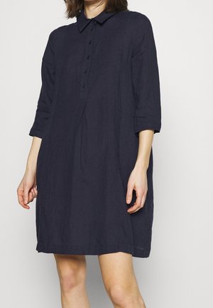 Kobieta ubrana w luźną, sięgającą kolan granatową sukienkę z rękawami do połowy przedramienia i koszulowym kołnierzykiem na guziki.