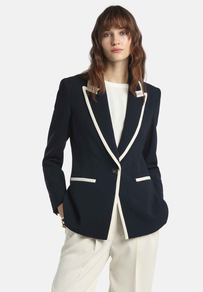 Námornícky modrý blejzer s bielym lemovaním, zapínaním na jeden gombík a dvoma prednými vreckami; kombinovaný s bielym topom a krémovými nohavicami. Hladká látka.