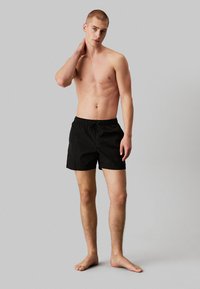 Pantaloni da nuoto neri con vita elastica e cordino. Leggeri, dalla texture liscia, di lunghezza al ginocchio. Il modello è in piedi a piedi nudi su uno sfondo neutro.