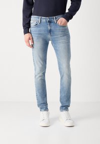 Jeans de mezclilla azul claro con un corte slim, que presentan un ligero desgaste, diseño de cinco bolsillos y cierre de botón, combinados con zapatillas blancas.