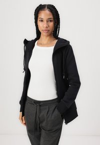 Haut à zip noir avec cordons de serrage, porté sur un débardeur blanc. Associé à un pantalon de survêtement gris avec cordon de serrage. Tissu lisse, design décontracté.
