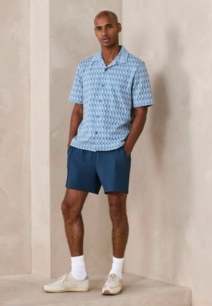 Man wearing a blue patterned short-sleeve button-up shirt, navy shorts, white socks, and beige sneakers, standing with hands in pockets.