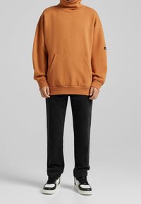 Sudadera naranja de gran tamaño con cuello alto, bolsillo frontal y dobladillo acanalado, combinada con pantalones vaqueros negros de pierna recta y zapatillas en blanco y negro.