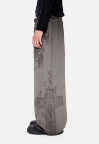 Άτομο ντυμένο με γκρι παντελόνι φαρδιάς γραμμής με μαύρη περίπλοκη σταυρωτή κέντηση, μαύρα παπούτσια και μαύρο μακρυμάνικο τοπ, που στέκεται πλευρικά.