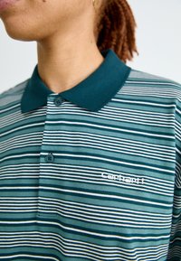 Polo a righe in teal, verde scuro e bianco. Presenta un colletto con bottoni, maniche corte e un piccolo logo ricamato sul petto.