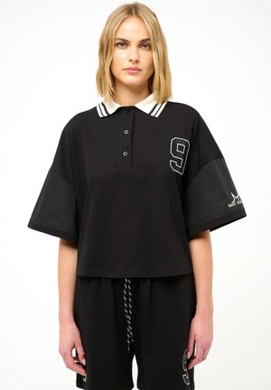 Vrouw draagt een losse zwarte polo met korte mouwen, een witte kraag en een patch met het cijfer negen, gecombineerd met zwarte short met trekkoord en nummer negen.