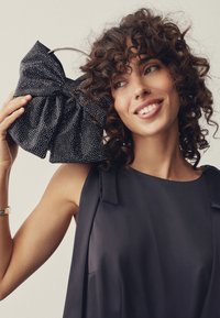 Juoda rankinė su lankelio dizainu, puošta sidabriniais smeigtukais. Turi metalinę apvalią rankeną. Modelis dėvi tamsią be rankovių palaidin�ę.