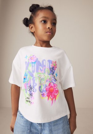 T-shirt bianco a maniche corte con grafiche colorate di fiori e farfalle accompagnate da testo, abbinato a pantaloni corti in denim blu chiaro.