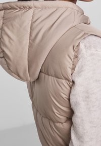 Beige zateplená vesta s kapucí, s prošívanou texturou a hladkým povrchem. Detail látky na rameni s jemným bílým pleteným trikem pod ním.