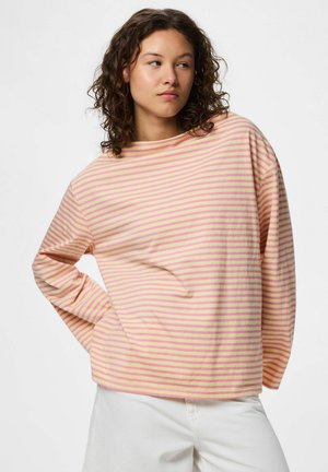 Ung kvinde med krøllet hår bærer en gul og pink stribet langærmet skjorte og hvide bukser, stående med den ene hånd på hoften.