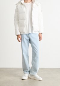 Mężczyzna w białej kurtce puchowej, białym t-shircie, jasnoniebieskich dżinsach i białych sneakersach stoi na szarym podłodze przy białej ścianie.