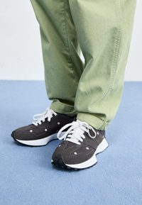 Mørkegrå sneakers med hvite snørebånd og blomsterdetaljer, med en tykk hvit såle og teksturert materiale. Brukt med lysegrønne bukser.