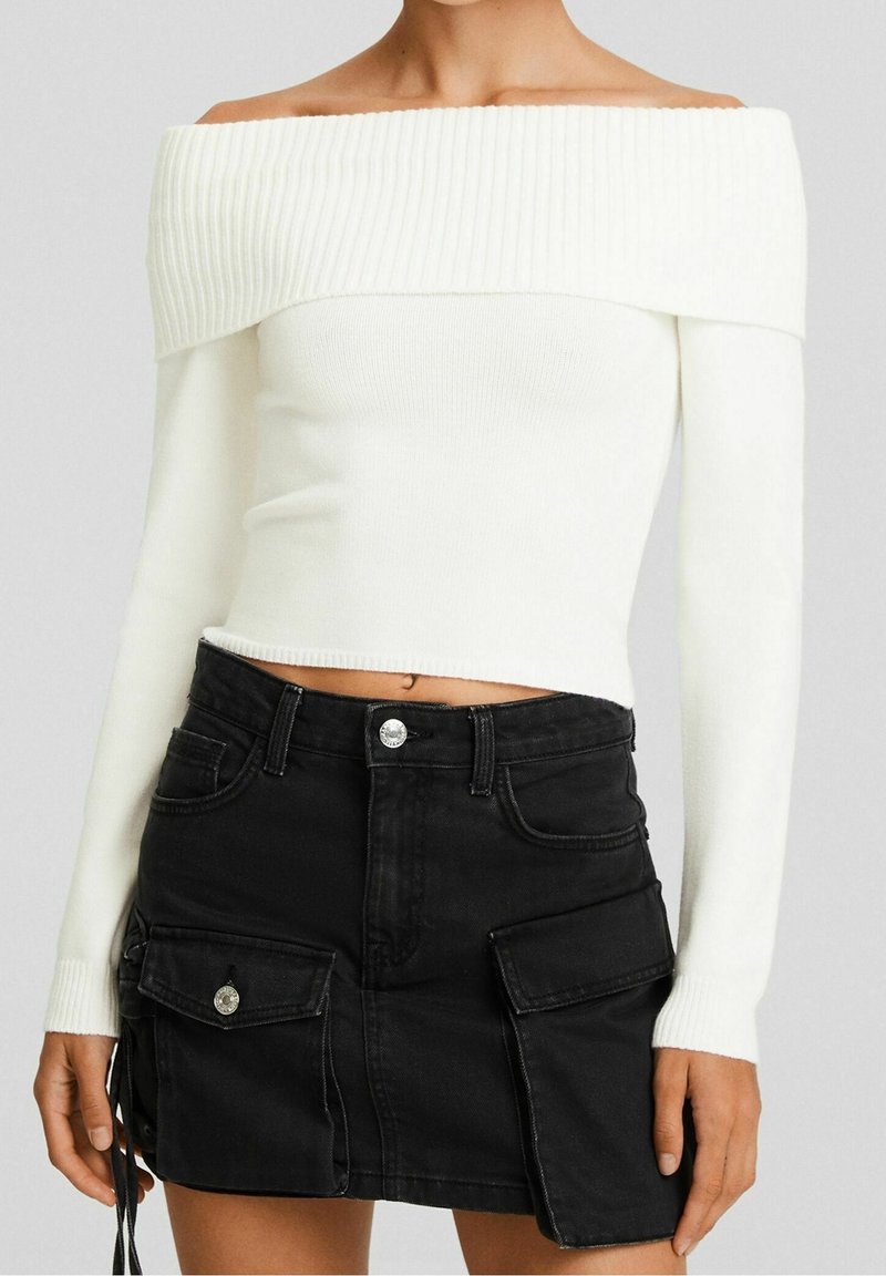 Femme portant un pull blanc côtelé décolleté aux épaules et une mini-jupe en jean noire boutonnée avec des poches cargo.