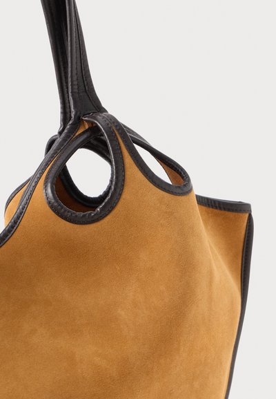 Suede tote bag in tan with black leather trim. Features two circular handles and a soft texture, emphasizing its structured yet relaxed form.