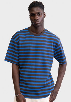 Giovane uomo con capelli corti attorcigliati che indossa una maglietta a maniche corte a strisce orizzontali blu e marroni, in piedi contro uno sfondo chiaro e uniforme.