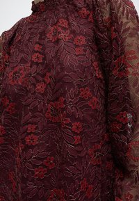 Δαντέλα βουργουνδί με περίπλοκο λουλουδένιο μοτίβο σε κόκκινους και σκούρους τόνους, με διαφανή μανίκια και λεπτομέρειες με σούρες στον λαιμό.