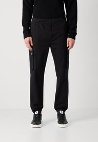 Zwarte cargobroek met een elastische tailleband, steekzakken en omgeslagen enkels, gemaakt van een gladde, duurzame stof. Gepaard met zwarte sneakers.