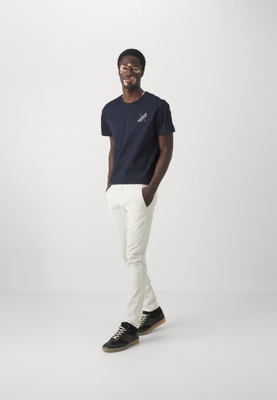T-shirt di cotone navy con logo, abbinata a pantaloni bianchi slim-fit e sneakers nere con lacci crema e suola in gomma.