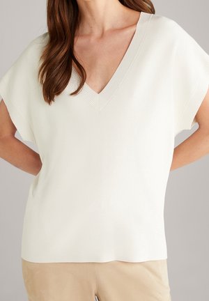 Femme portant un haut en maille blanc ample à manches courtes et col en V avec des bords côtelés, et un pantalon beige sur un fond uni.