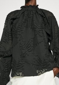 Blouse noire transparente avec motifs floraux texturés, col montant volanté et manches longues bouffantes, portée sur un pantalon blanc.