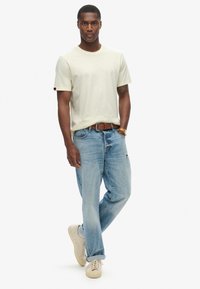 Lys beige T-shirt med korte ærmer, parret med lys blå distressed jeans. Model iført casual beige sneakers og et brunt bælte.