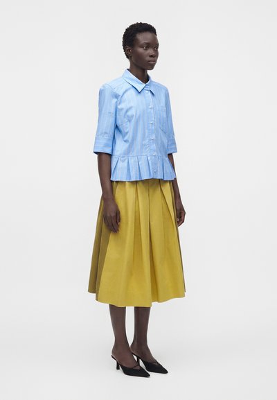 Femme portant un chemisier bleu clair rayé à boutons avec manches aux coudes et bas à basque, associé à une jupe midi plissée jaune et des talons noirs.