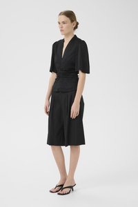 Black short-sleeve top with a gathered waist, paired with black knee-length shorts. Model wears black heeled sandals. Plain white background.