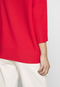 Maglione rosso a maglia con finitura testurizzata, caratterizzato da un collo a barca e maniche a tre quarti, abbinato a pantaloni bianchi a gamba larga.